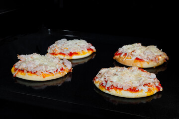 Four baked mini pizzas with ham, cheese and sause on tray in electric oven. Italian cuisine, homemade bakery, fast food, cooking concept - obrazy, fototapety, plakaty