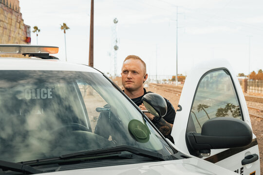 Horizontal image of white male caucasian police officer standing in the door opening of his trooper car with lights on looking away from camera next to railroad tracks.