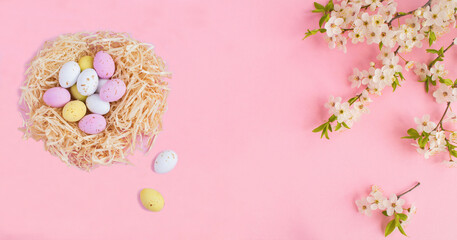 Fototapeta na wymiar Colored easter eggs in a nest of straw and white flowering tree branches on the pink background. Holiday background. Copy space.