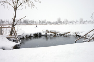 Winter landscape. A frozen river covered with snow with an ice hole in the foreground. The hole is equipped with a ladder and . In the distance, a lone fisherman with a fishing rod at the hole.