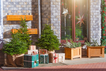 Three freshly live fir trees and boxes with gifts near the building decorated with decorations lit by the bright sun.