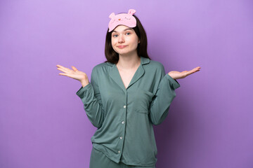 Young Russian woman in pajamas isolated on purple background having doubts while raising hands