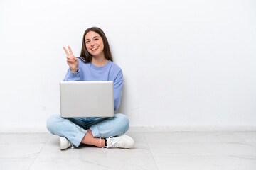 Young caucasian woman with laptop sitting on the floor isolated on white background smiling and showing victory sign