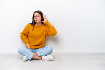 Young girl sitting on the floor isolated on white background listening to something by putting hand...