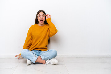 Young girl sitting on the floor isolated on white background with glasses and surprised