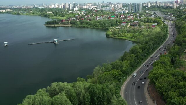 Modern highway with driving cars near tranquil lake against large city on summer day bird eye view. Picturesque riverside