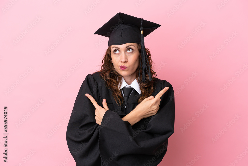 Canvas Prints Young university graduate woman isolated on pink background pointing to the laterals having doubts