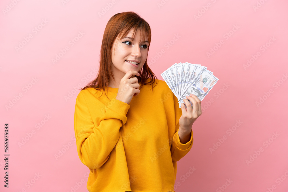 Wall mural Redhead girl taking a lot of money isolated on pink background looking to the side and smiling