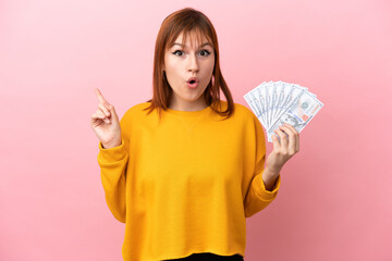 Redhead girl taking a lot of money isolated on pink background intending to realizes the solution while lifting a finger up