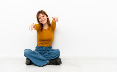 Redhead girl sitting on the floor isolated on white background points finger at you while smiling