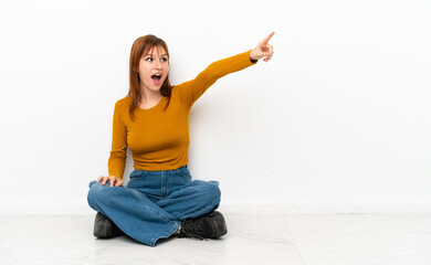 Redhead girl sitting on the floor isolated on white background pointing away