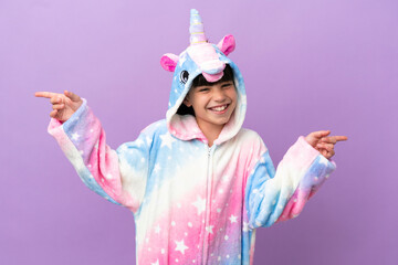 Little kid wearing a unicorn pajama isolated on purple background pointing finger to the laterals and happy