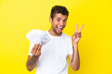 Young brazilian man taking a lot of money isolated on yellow background smiling and showing victory sign