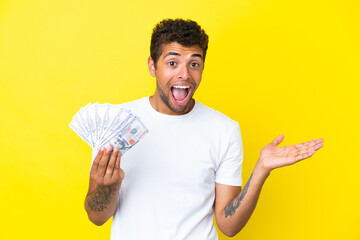 Young brazilian man taking a lot of money isolated on yellow background with shocked facial expression