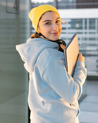 Smiling female student in a blue sweatshirt and yellow hat is walking down the corridor with a...