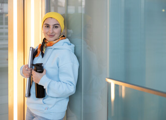 A girl in a blue sweatshirt and yellow hat stands in the corridor and holds a computer and a thermos in her hands.