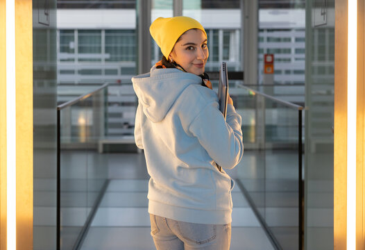A smiling female student in a blue sweatshirt and yellow cap walks along the glass corridor 