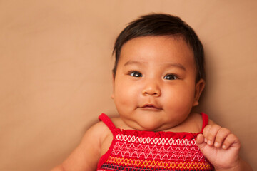Close-up portrait of Asian baby lying on back, wearing red dress, looking at camera and smiling, studio shot on brown background