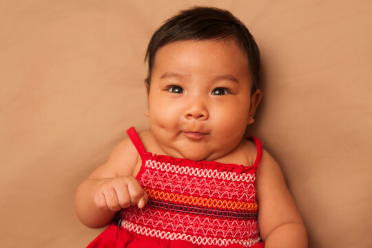 Close-up portrait of Asian baby lying on back, wearing red dress, looking at camera and making funny face, studio shot on brown background