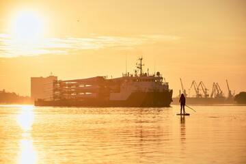 Obraz premium Girl standing on paddle board near cargo vessel loaded with rotor blades for wind turbines at the sunrise Danube river