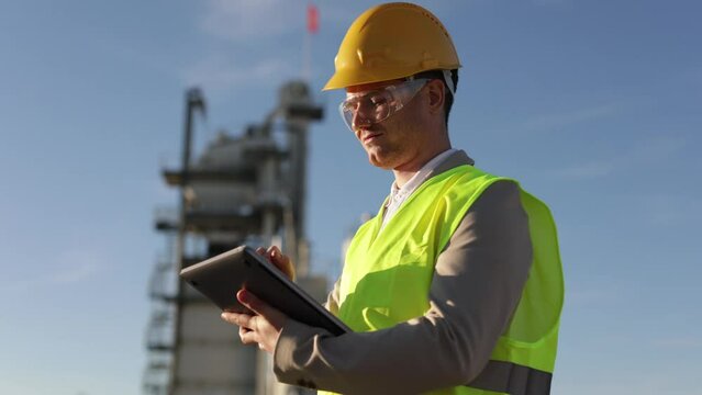 A professional engineer, a heavy industry worker, an inspector dressed in a protective uniform and a helmet works with a tablet computer against the background of a huge factory.