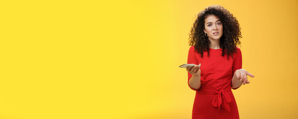 So waht, I confused. Questioned uncertain woman with curly hair in red dress shrugging looking clueless as holding hand cannot understand where order as checking mail box via device over yellow wall - obrazy, fototapety, plakaty