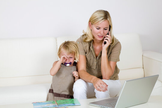 Mother Using Laptop and Cell Phone, Toddler Standing Beside Her Crying