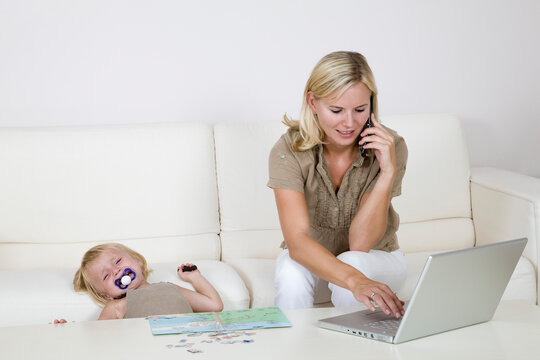 Mother Using Laptop and Cell Phone, Toddler Sitting Beside Her Crying