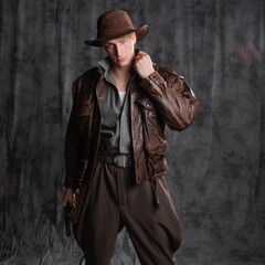 A young man in retro style, an adventure character. The hero of the adventure, a guy in a hat and a leather jacket, posing in the studio on a gray background - obrazy, fototapety, plakaty