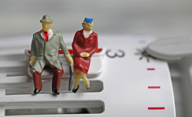 Obraz premium Figures of old retired couple sitting warmly dressed on a heater thermostat at cold temperatures on saving clip - Pensioners poor people heating costs concept