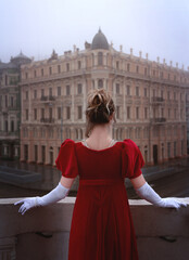 Beautiful young girl in a nineteenth century dress stands on a balcony in front of a beautiful building.