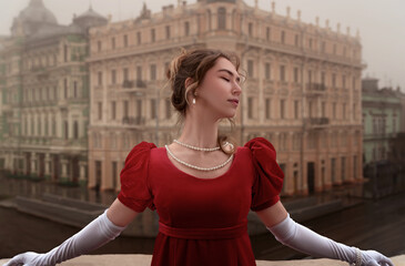 Beautiful young girl in a nineteenth century dress stands on a balcony in front of a beautiful building. - obrazy, fototapety, plakaty