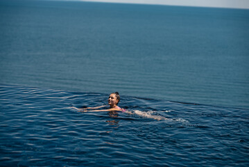 Happy woman in swimsuit swimming in infinity pool against seafront.