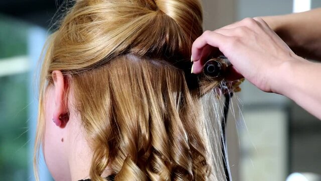 Hands of professional stylist twist blonde strands using curling iron on customer hair. Client sits in chair waiting for modern hairstyle close backside view