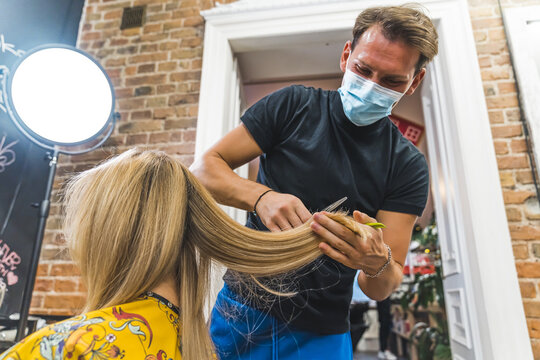 Hairstyling during pandemic. Professional male hairstylist in protective face mask cutting the ends of blond hair. Hair salon interior. High quality photo