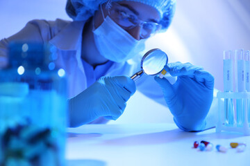 Scientist with magnifying glass examining pill at table in laboratory, closeup
