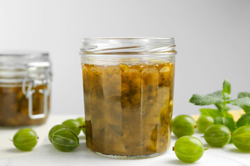 Jars of delicious gooseberry jam and fresh berries on white table, closeup