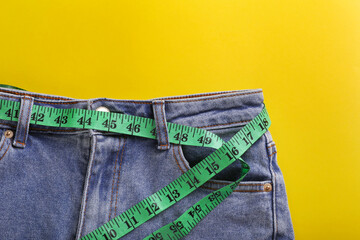 Jeans with measuring tape on yellow background, top view. Weight loss concept