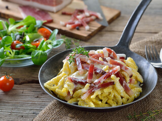 Deftige Spätzle mit Käsesauce und Südtiroler Bauernspeck in der Eisenpfanne mit Salatbeilage serviert - Hearty spaetzle with cheese sauce and South Tyrolean bacon served in an iron frying pan