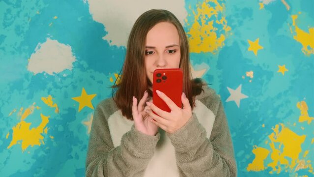 Portrait of beautiful woman in plush hoodie browsing mobile phone, smiling, on background of bright wall.