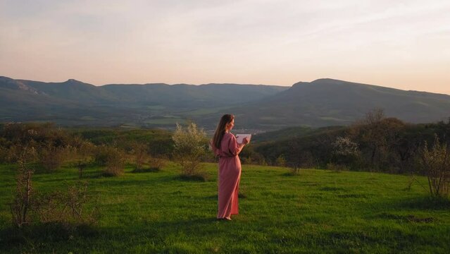 the girl stands against the background of a beautiful landscape at sunset and draws. a girl paints at sunset. the artist paints a landscape. En plein air painting. sketch of the landscape