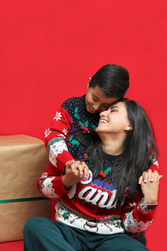 Latina Mom And Son Wear Ugly Christmas Sweaters And Show Their Love To Each Other On A Red Background Between Large Gift Boxes With Bow