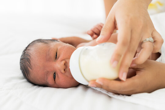 Hands of mother or nurse feeding newborn baby with milk bottle. Newborn born sleeping and eating milk from milk bottle nipples on bed