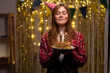 Happy 18 years old girl in party cone make a wish and blowing out candles on birthday cake,...