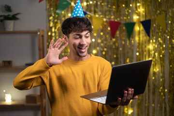 Virtual celebration. Portrait of happy young arabic man wearing party hat holding laptop and waving...