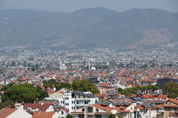 Fototapeta na wymiar A view of the city from above against a background of towering mountains and hills. Panorama of the Turkish city of Fethiye with mosques and minarets. Tiled roofs of houses with solar panels