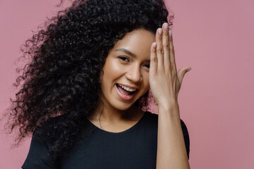 Happy teenage girl has fun and hides face with palm, has joyful expression, curly bushy hair,...