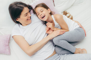 Photo of adorable small child and her mother have fun together in bed, tickle each other, smile...