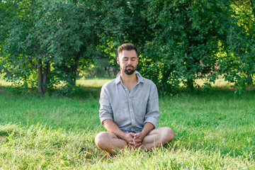 Portrait of young handsome European man in casual clothing sitting on a grass in summer park.