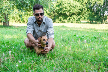 Naklejka na ściany i meble Handsome stylish European man sitting on grass with his dog in park on a walk. Friendship between human and pets.